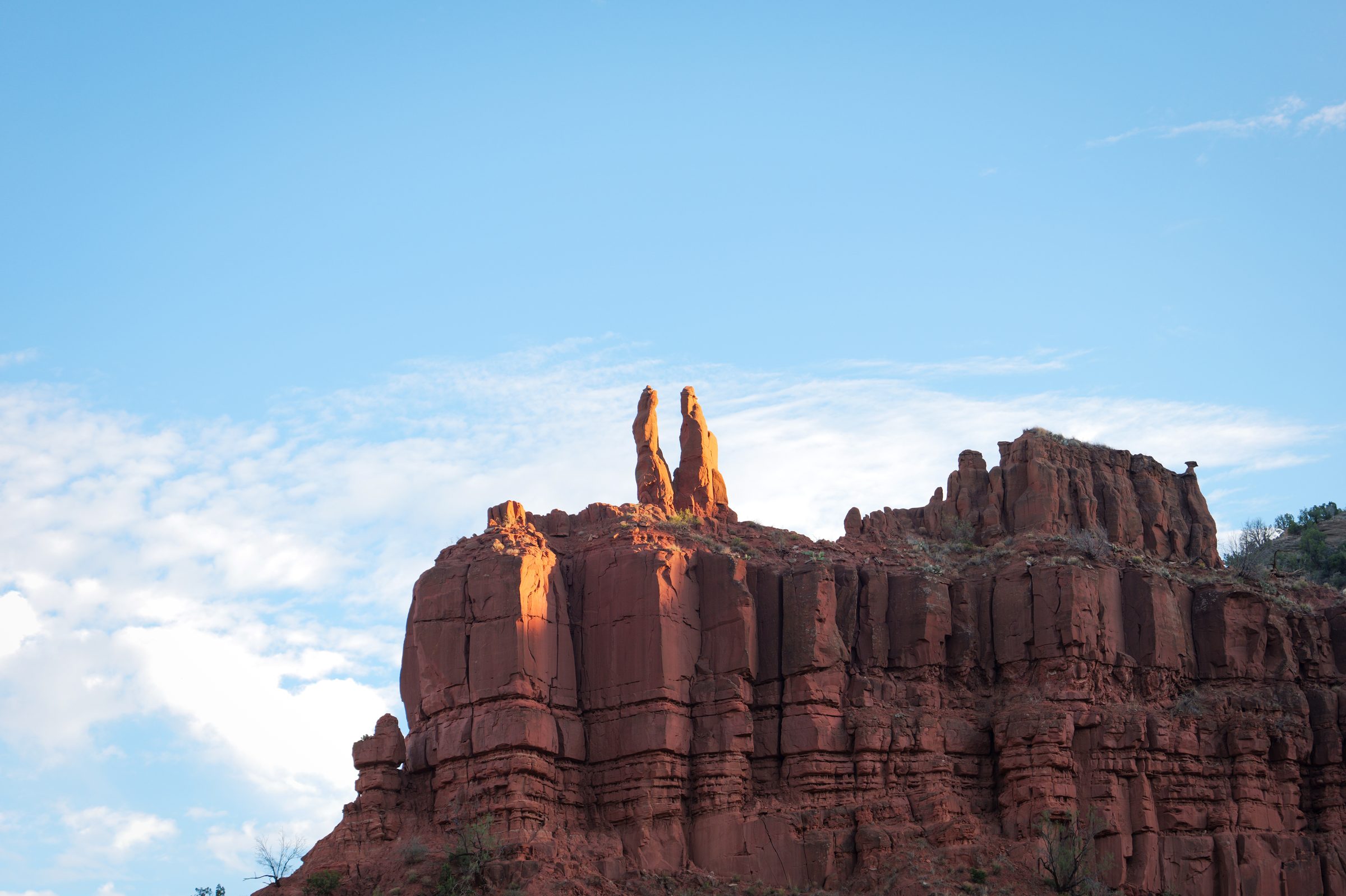 Muro de roca roja con dos agujas de piedra en la cima, iluminadas por la luz dorada de la tarde.