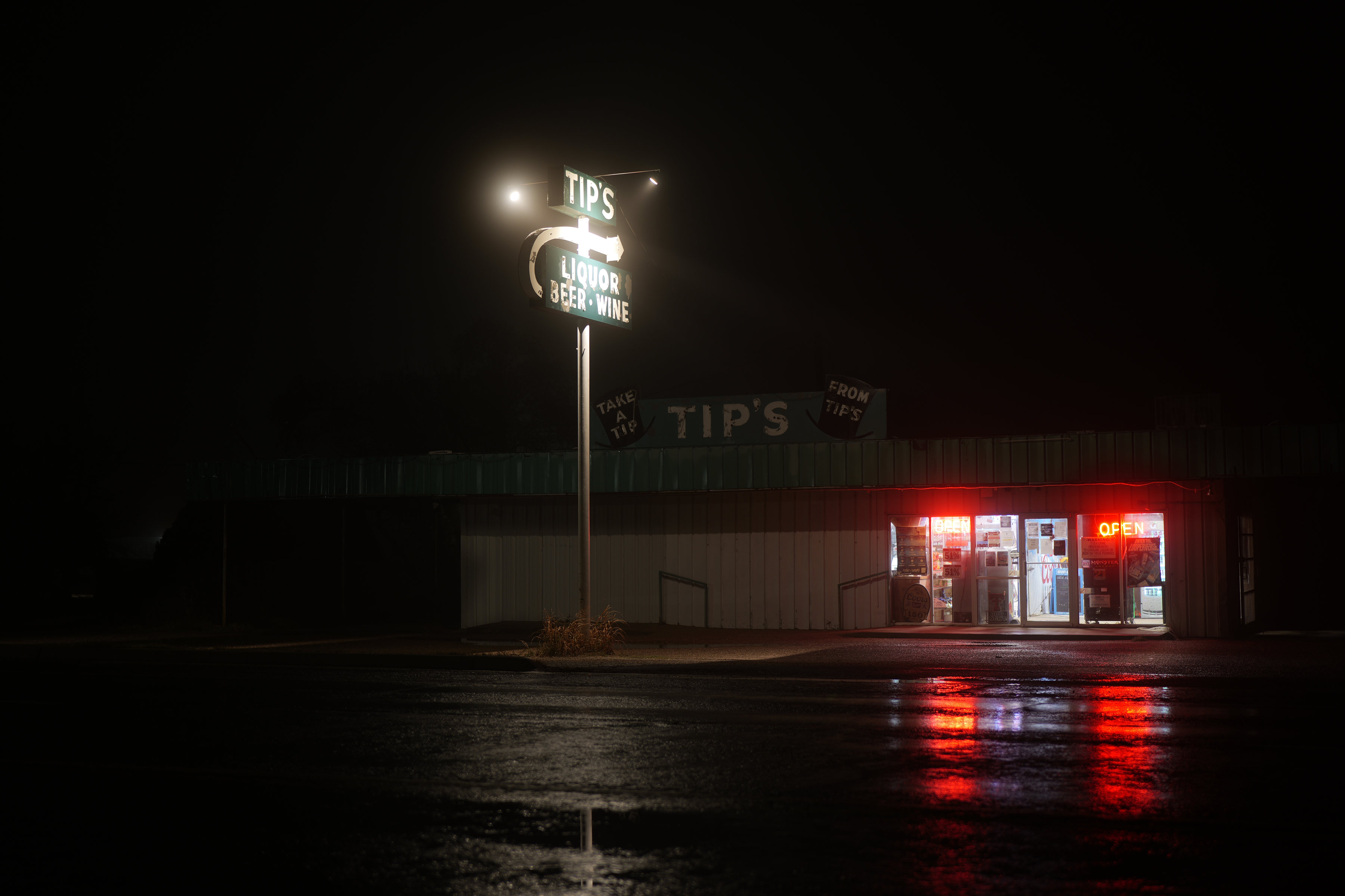 Letrero iluminado de una licorería de noche, con niebla y reflejos rojos de neón sobre el asfalto mojado.